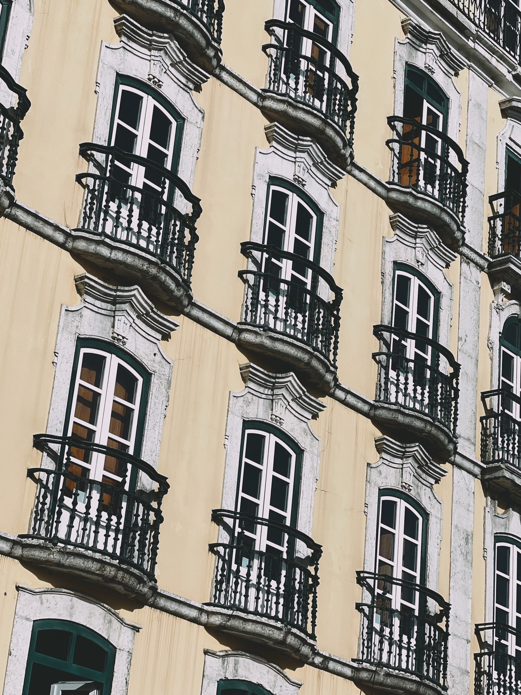 A building facade with rows of arched windows, each featuring ornate wrought iron balconies. The exterior is painted in a pale yellow with decorative stone frames around the windows.