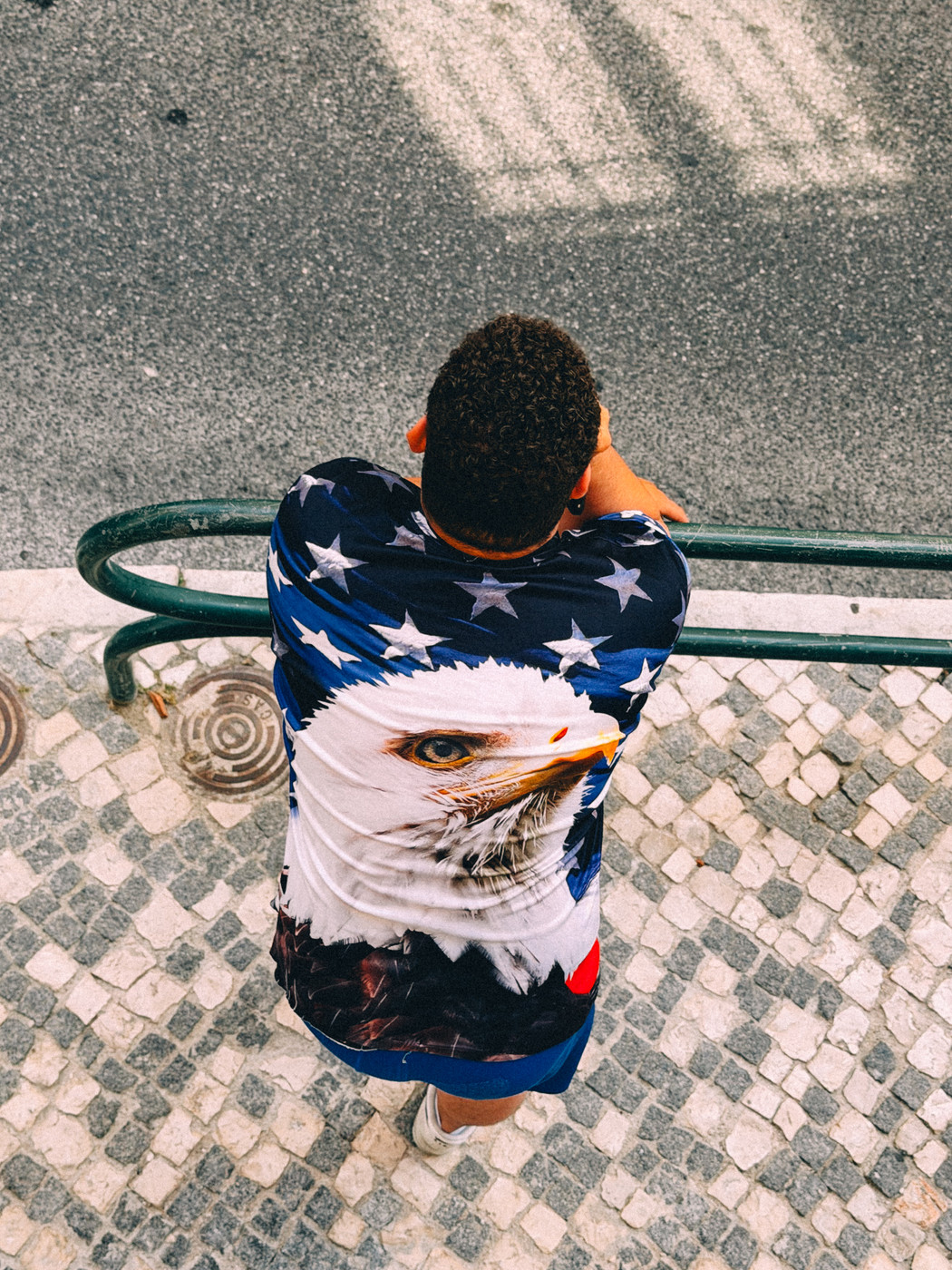 Person with short curly hair wearing a shirt featuring a bald eagle and white stars on a blue background, leaning on a green railing beside a cobblestone street.