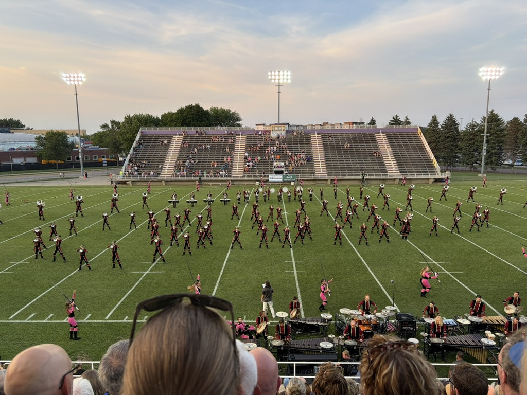The Madison Scouts Drum & Bugle Corps performs a field show in a stadium under the lights, with musicians spread across the football field, pit percussion at the front sideline, and spectators watching from the stands.