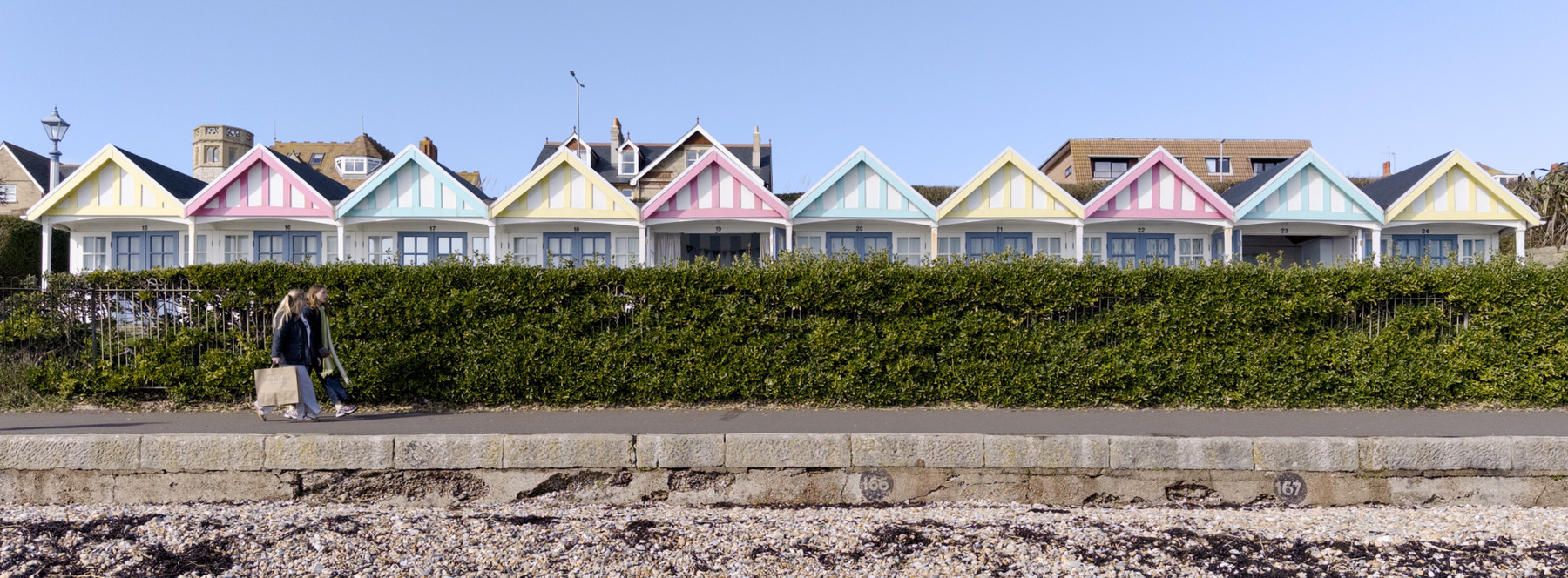 Two people walking along a path in front of a row of colourful beach huts. The huts are painted in pastel shades of yellow, pink, blue, and green, with white trim and gabled roofs. A tall hedge separates the path from the huts. The sky is clear and blue, suggesting a sunny day. The mood is calm and leisurely, evoking a sense of a pleasant seaside stroll.