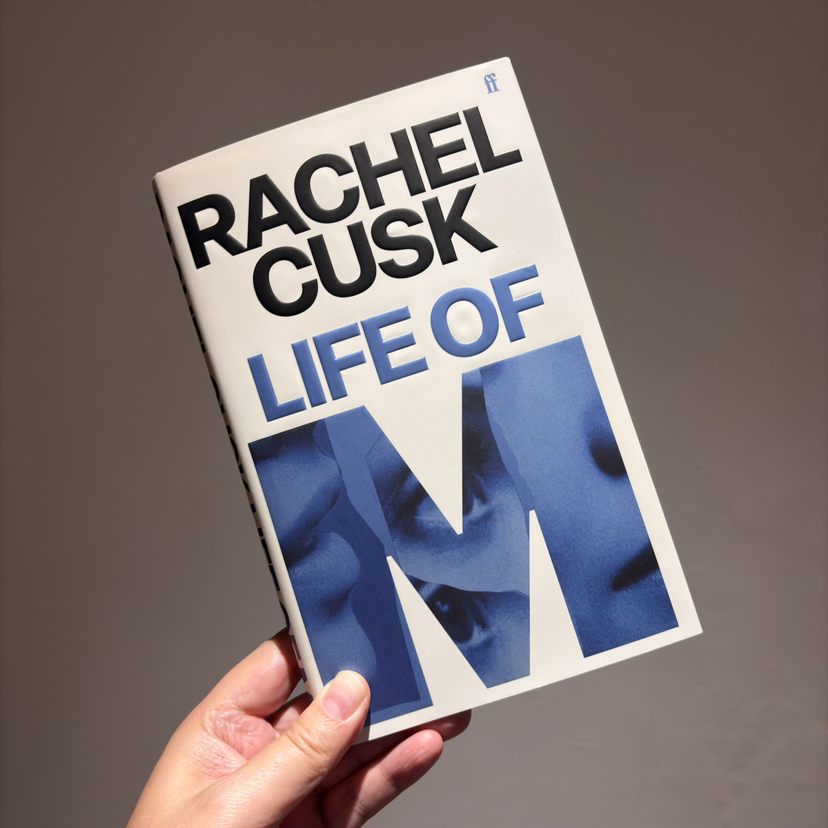 A hand holds up a hardback copy of ‘Life of M’ by Rachel Cusk. The title and author’s name appear in block capital letters on the cover.