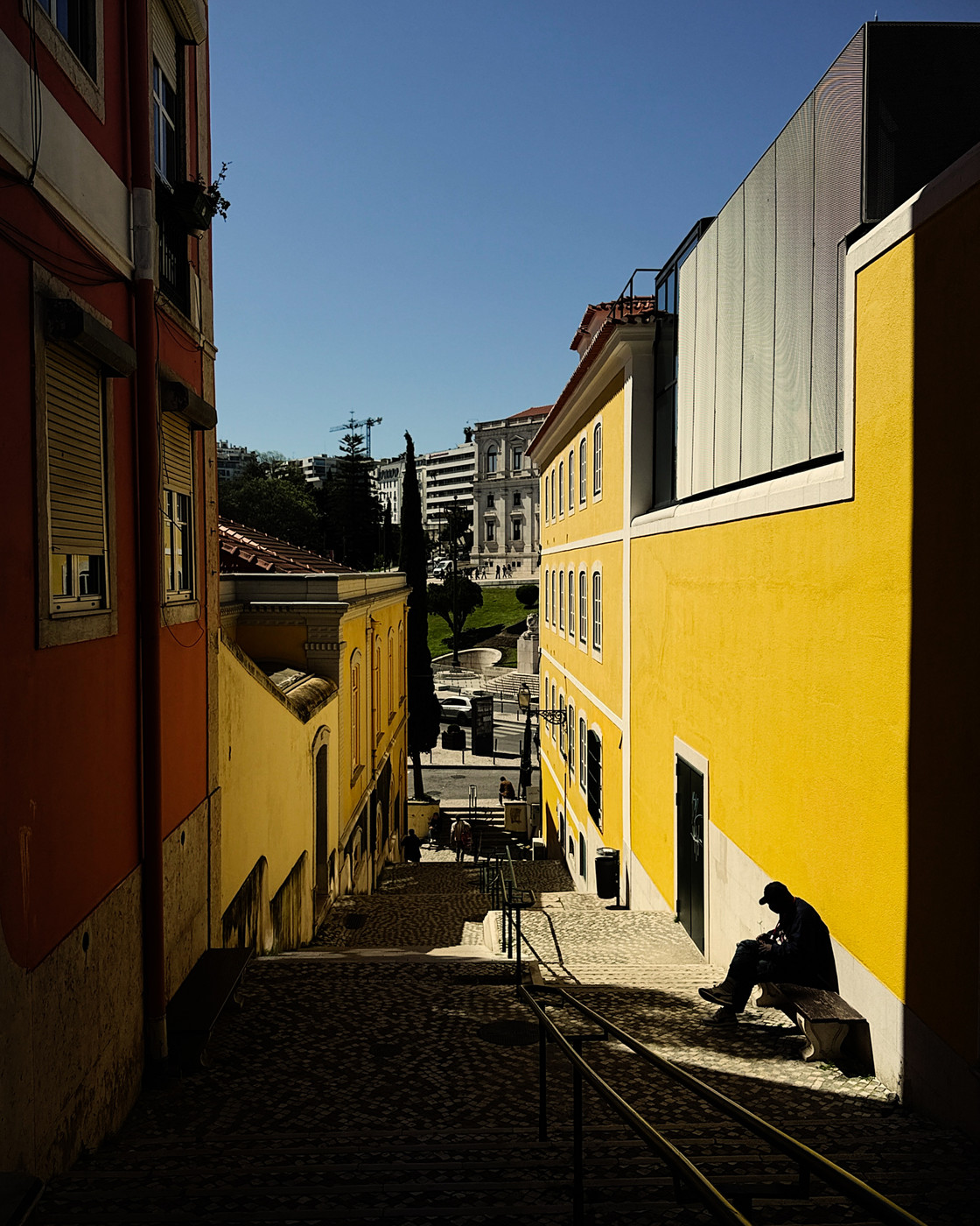 Narrow cobblestone alley with colorful yellow and orange buildings, a person sitting on a bench in the shadow, and a bright blue sky with distant modern and historic architecture.