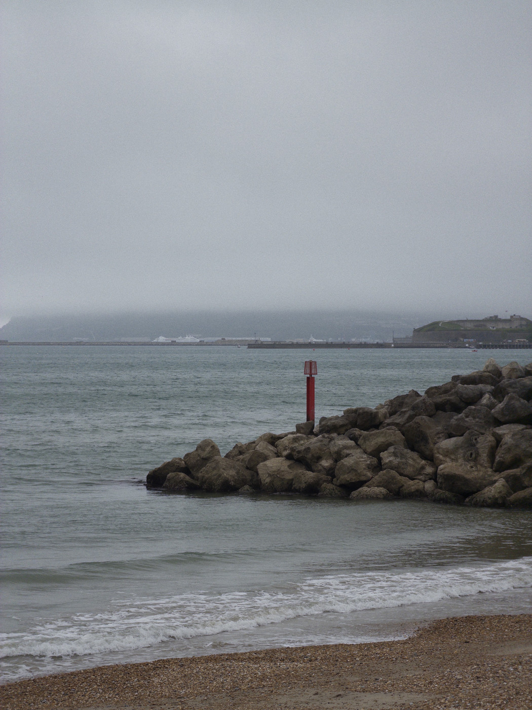 A grey, overcast coastal scene featuring a shingle and sand beach in the foreground, with gentle waves lapping the shore. A large rocky breakwater made of rough boulders extends into calm grey-green sea water, with a red cylindrical navigation marker mounted on top. In the middle distance, a harbour is visible with what appear to be large ferries or ships docked at a terminal. To the right, a headland rises from the water with a fortified structure on its summit. Misty hills or cliffs are partially obscured by low cloud in the background beneath a flat, overcast sky.