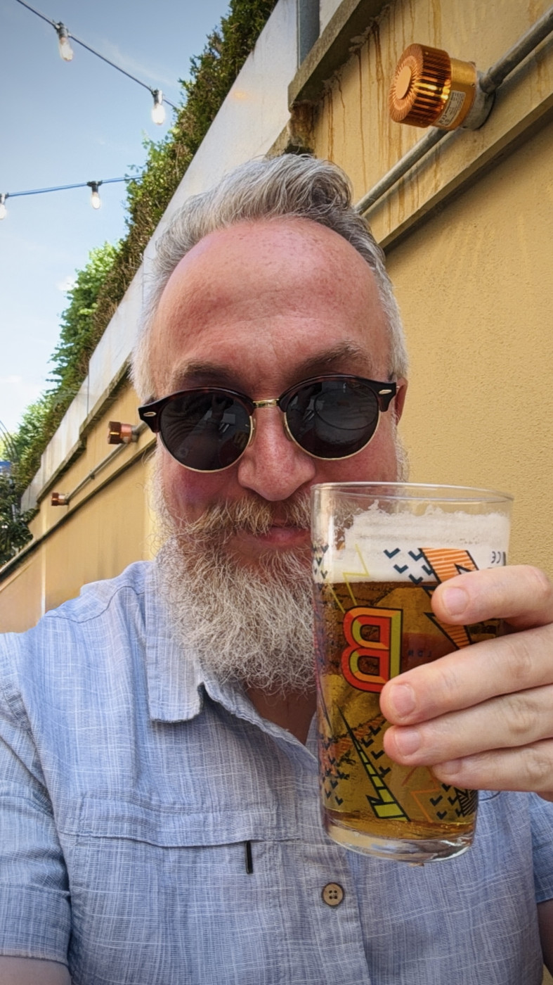 Grey-haired bearded man in sunglasses holding a pint of lager at a sunny pub garden with festoon lights.