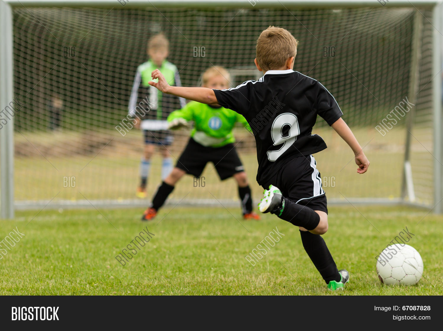 Stock photo of a young soccer player in a black jersey numbered 9 winding up to kick a soccer ball toward the goal, with a goalkeeper in a green jersey and a third player watching from behind the net on a grass field.