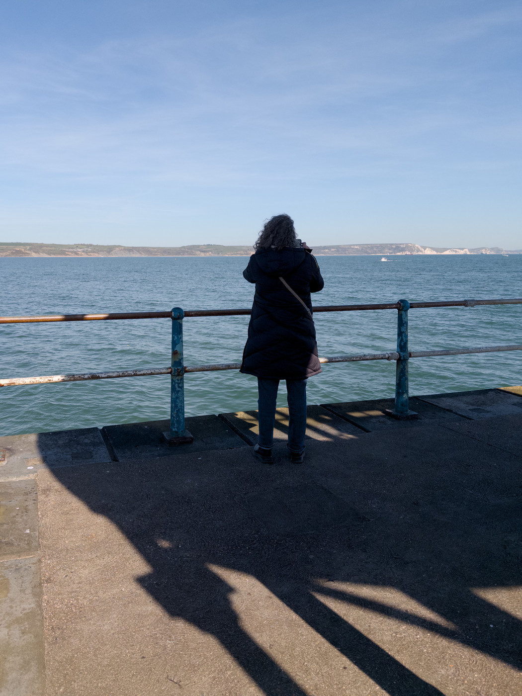 Person in a dark coat standing at a railing, looking out across the sea on a bright day.