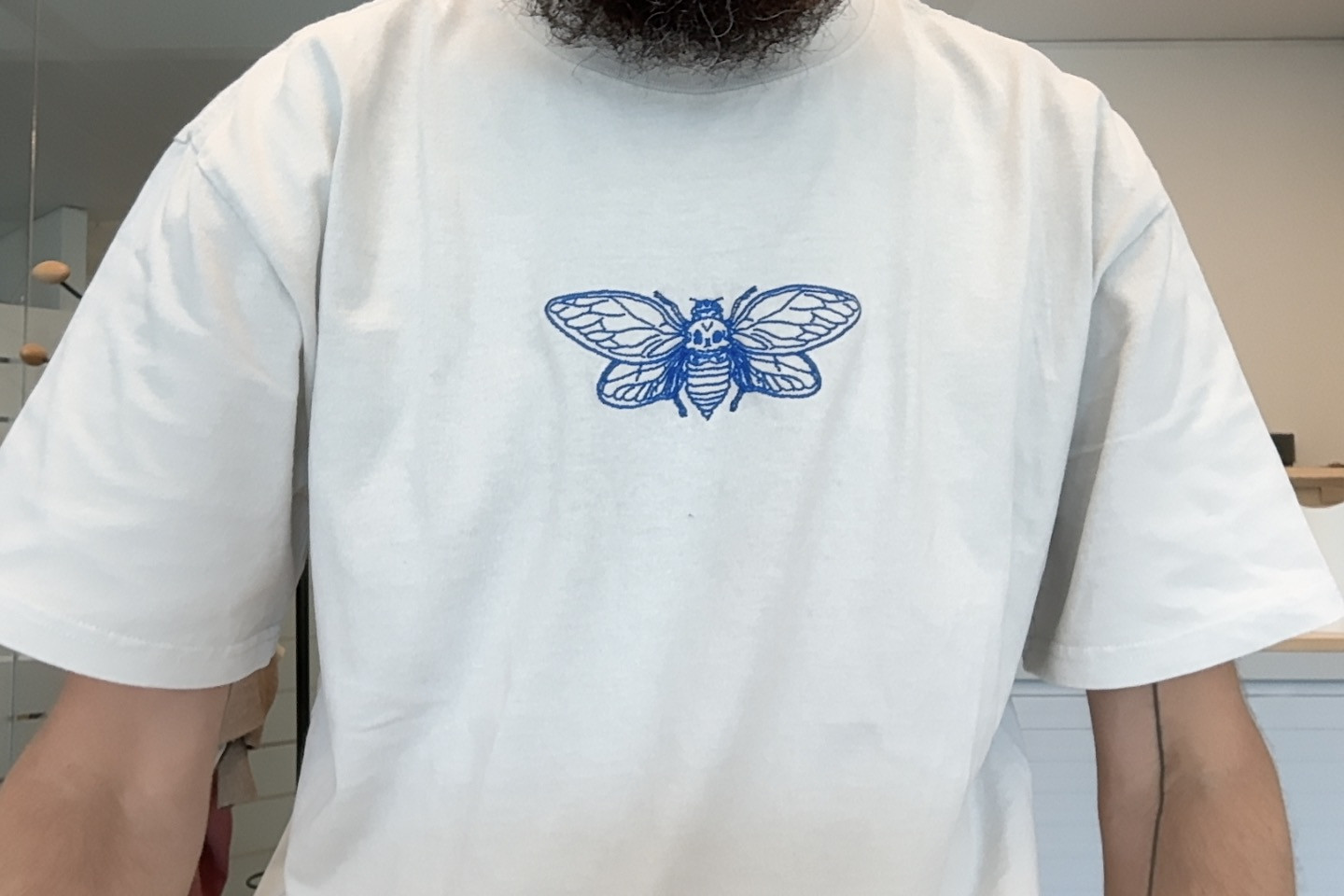 Close-up of a person wearing a plain white short-sleeve T-shirt with a blue, outlined moth graphic centered on the chest; the wearer has a beard and the image is cropped at the shoulders.