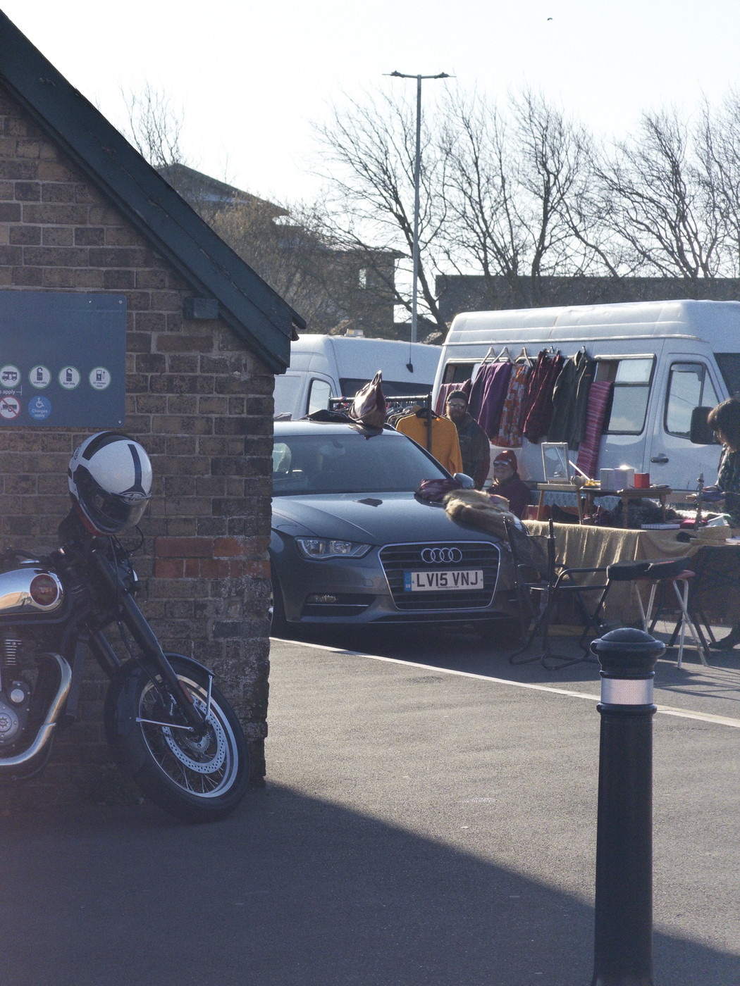 Street scene at an outdoor car boot sale on a sunny day. A grey Audi is parked beside stalls with clothes hanging from a van and tables of items, while a motorcycle with a helmet rests near a brick building in the foreground.
