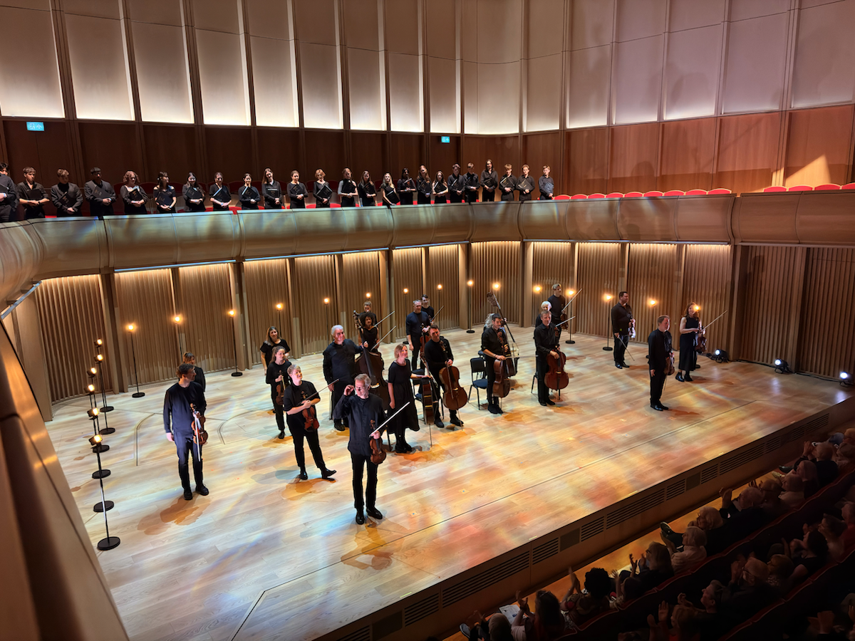 Orchestra standing on a wooden concert hall stage with string instruments, acknowledging applause from the audience, while a choir lines the balcony above.