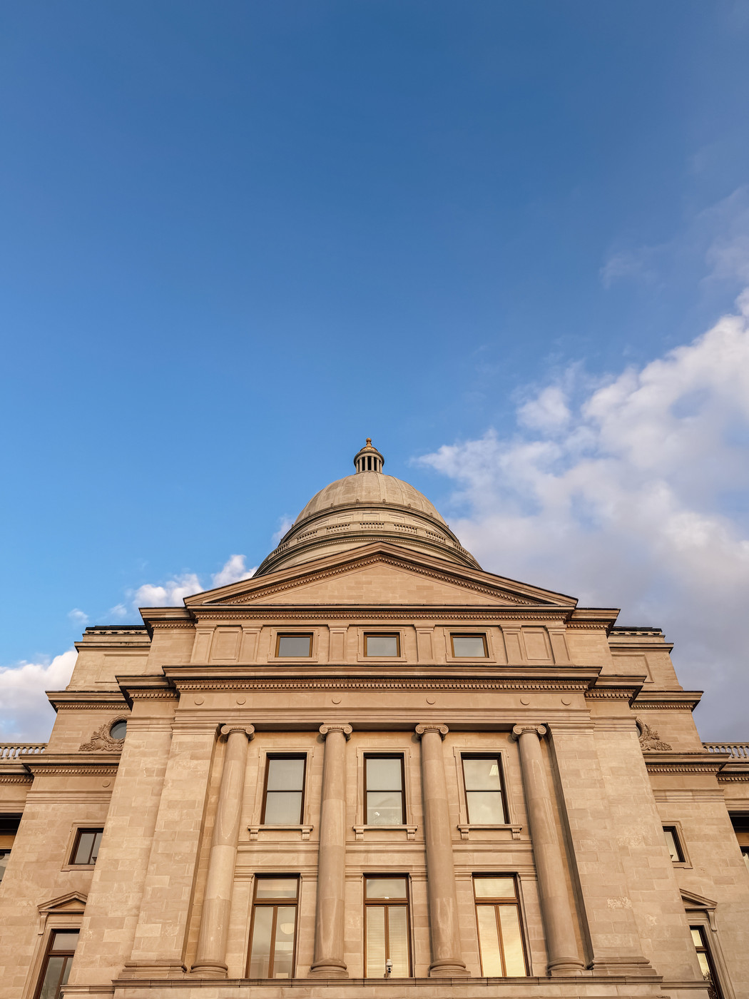 A neoclassical building with a dome and columns is set against a blue sky with scattered clouds.