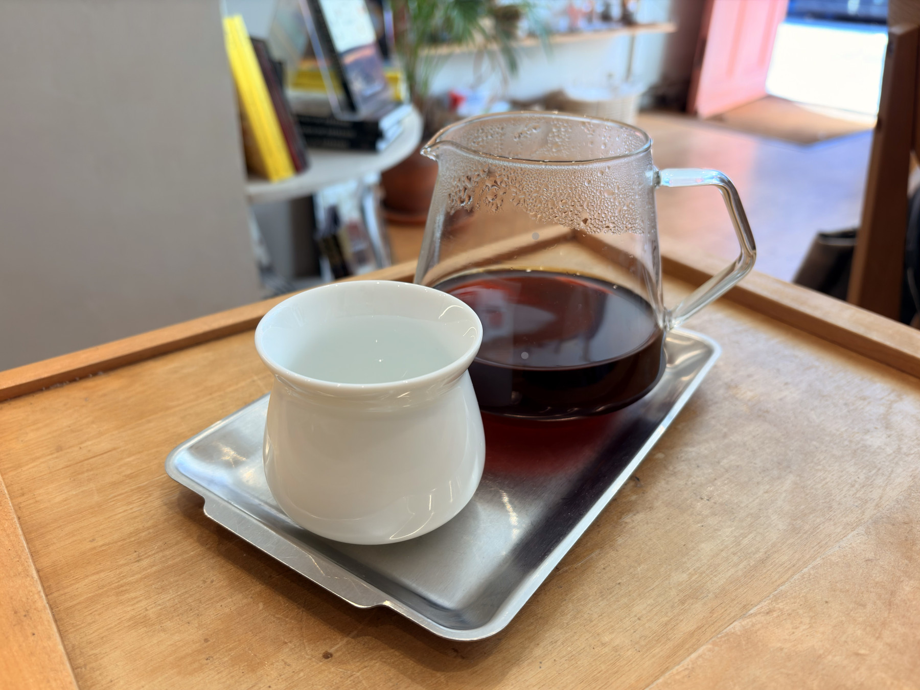 A glass carafe of coffee and a white ceramic drinking vessel on a metal tray sit on top of a wooden table.