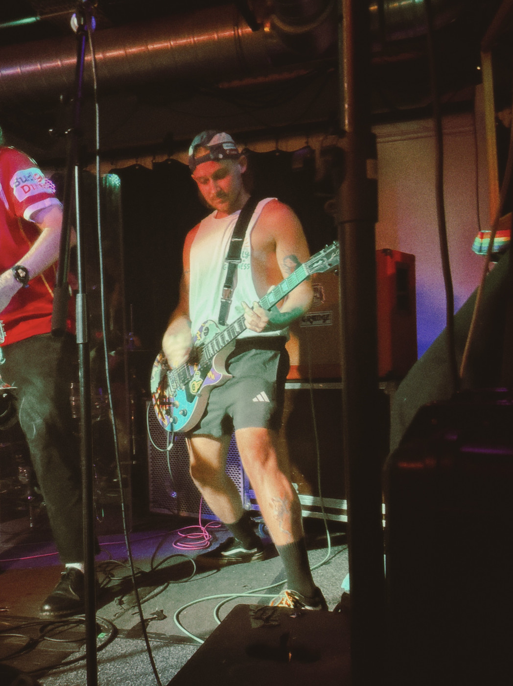 A male guitarist on a dimly lit stage wearing a white sleeveless shirt and a backward cap, playing a sticker-covered electric guitar. Cables lie on the floor and a microphone stand is in the foreground; another musician in red is partially visible to the left.