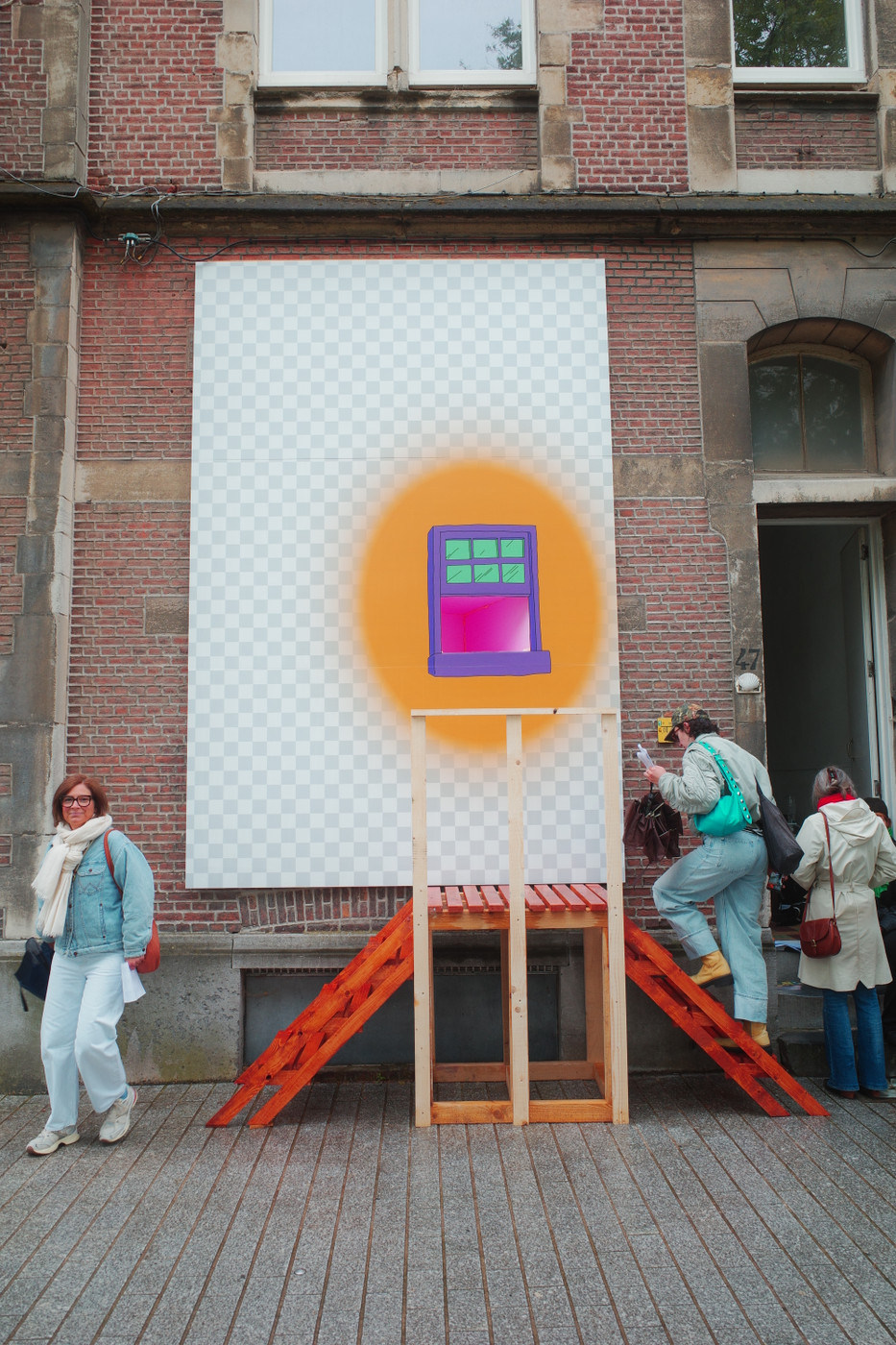 Outdoor street scene with a large mural on a red-brick building. The mural is a tall, white-and-light-gray checkerboard panel featuring a bright orange halo around a purple window with four green panes. A wooden scaffold with orange slats is in front of the artwork. Several pedestrians pass by: a woman in a light denim jacket and white scarf on the left, and others with bags on the right. Windows and stone architectural details are visible in the background.