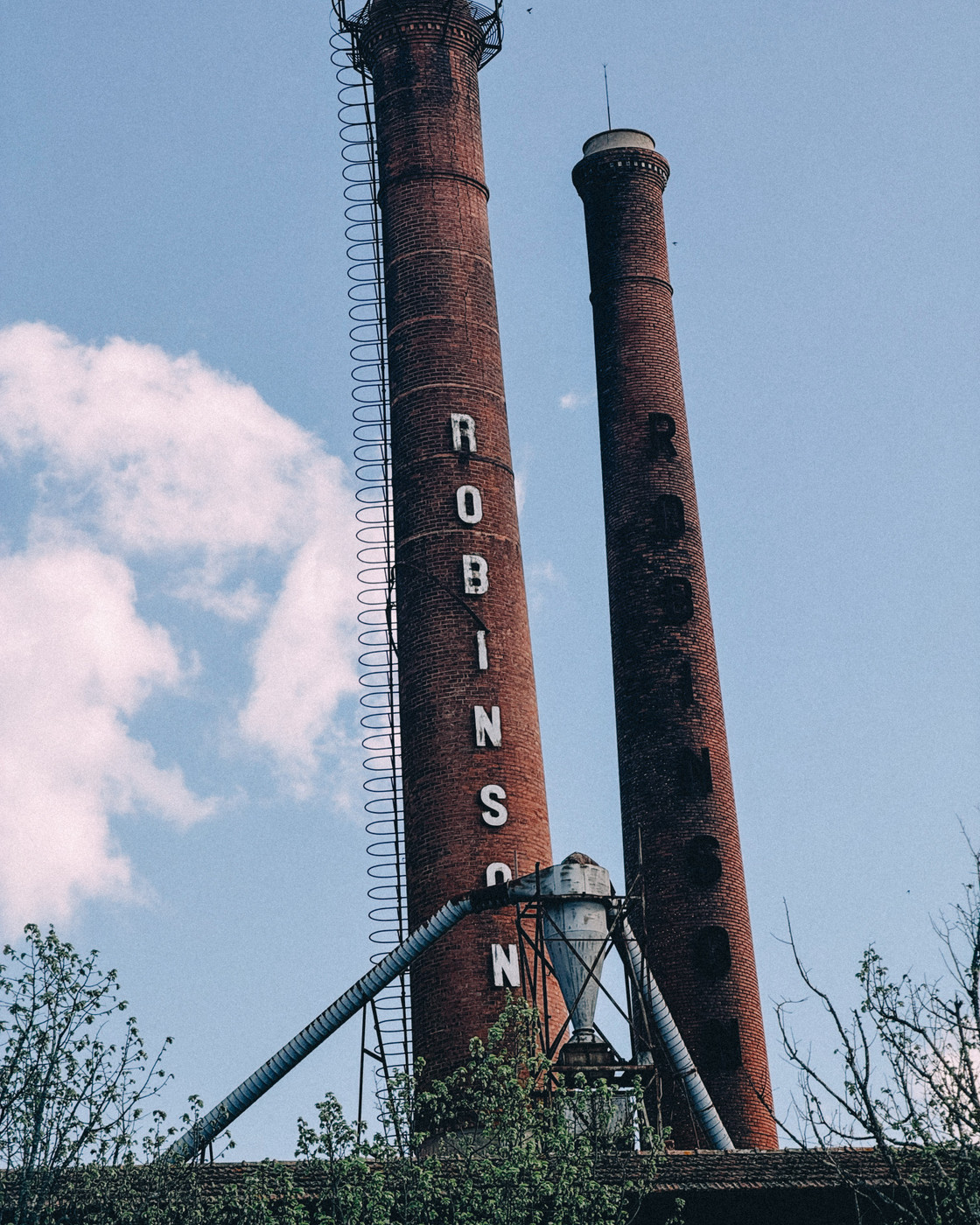 Two tall brick smokestacks with the word “ROBINSON” vertically displayed, surrounded by metal ladders and pipes, against a blue sky with scattered clouds and green tree tops.