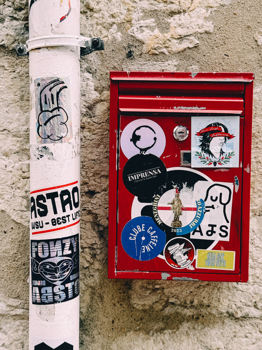 Red metal mailbox covered with various colorful stickers next to a white pole with graffiti and stickers, mounted on a textured stone wall.