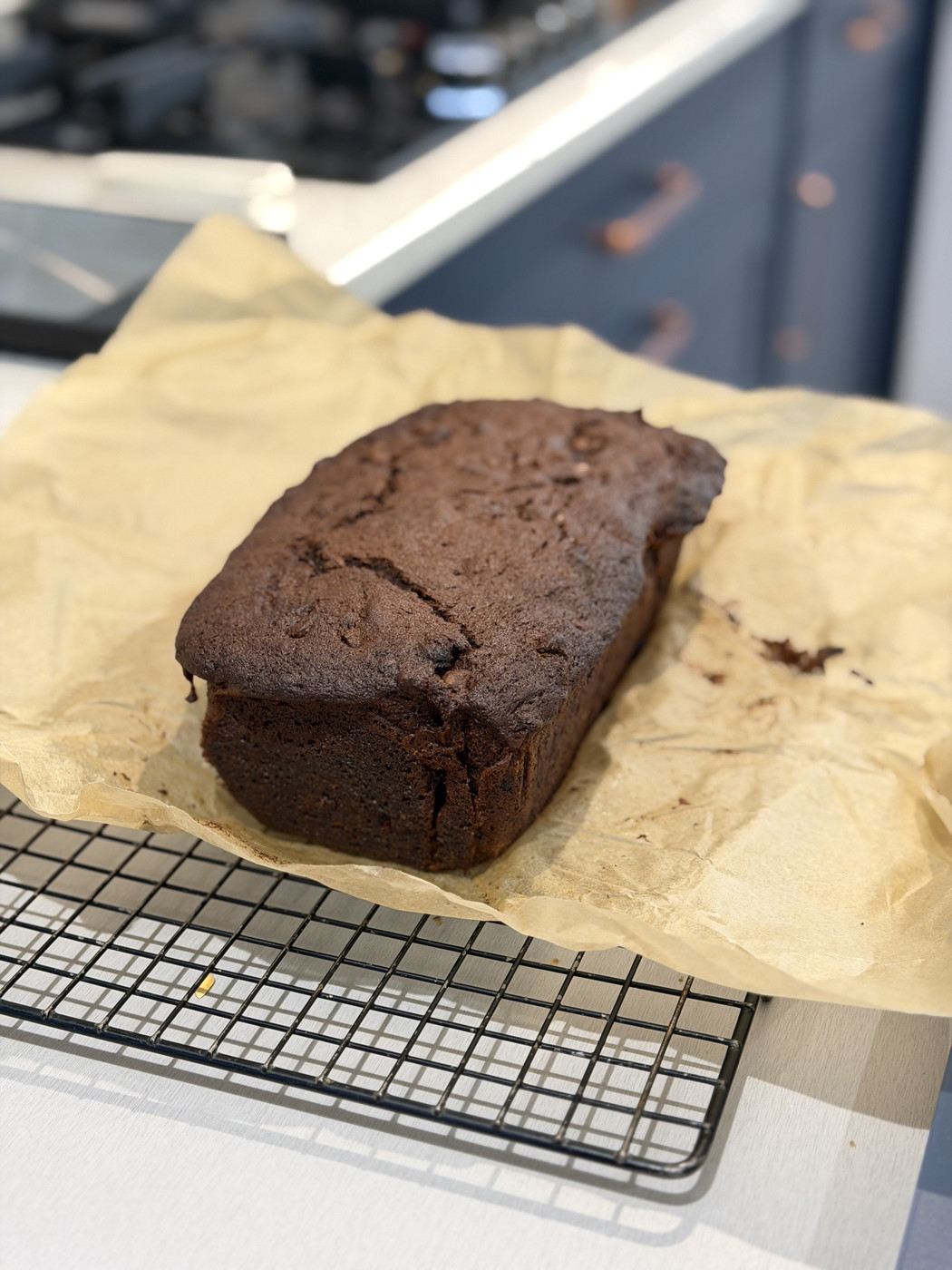 A dark brown cake loaf resting on crumpled parchment paper placed on a wire cooling rack. The background is part of kitchen with blue cabinets slightly out of focus.