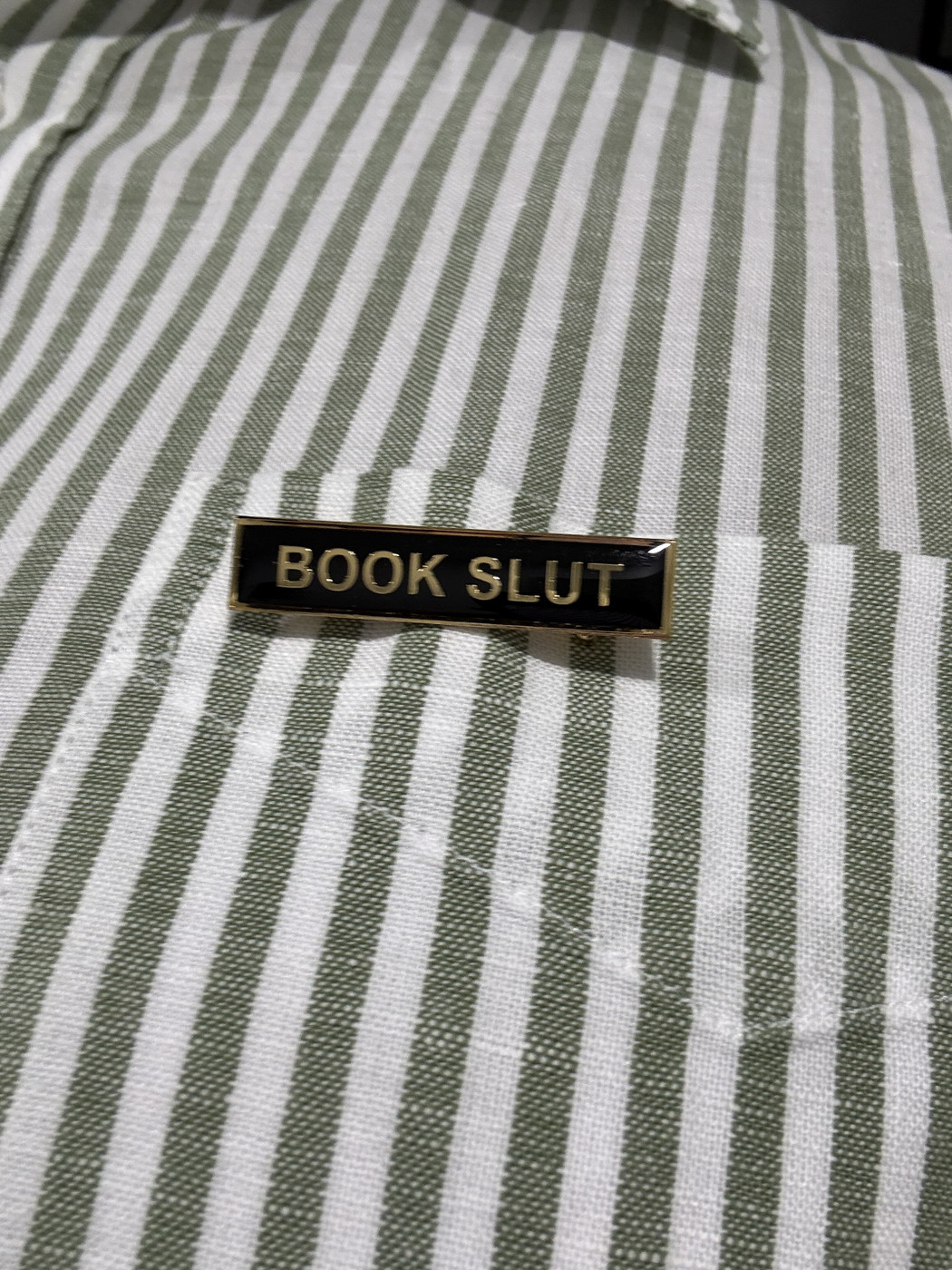 A rectangular enamel badge pinned on a green and white striped shirt. The badge has a black background and in gold all caps it says - Book Slut