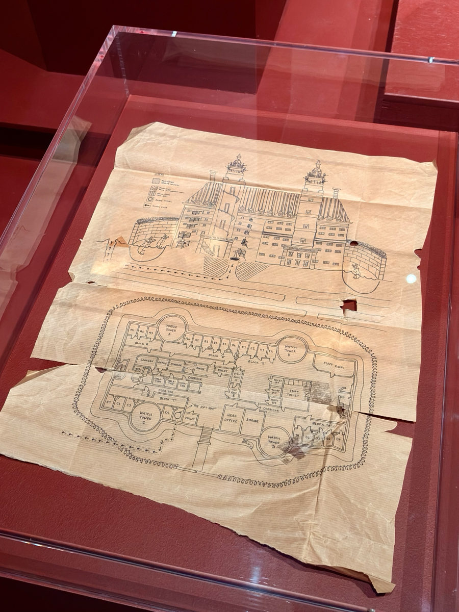Glass case displaying a fragile, yellowed paper plan with hand-drawn architectural elevations and floor layouts of a large institutional building, set against a deep red background.