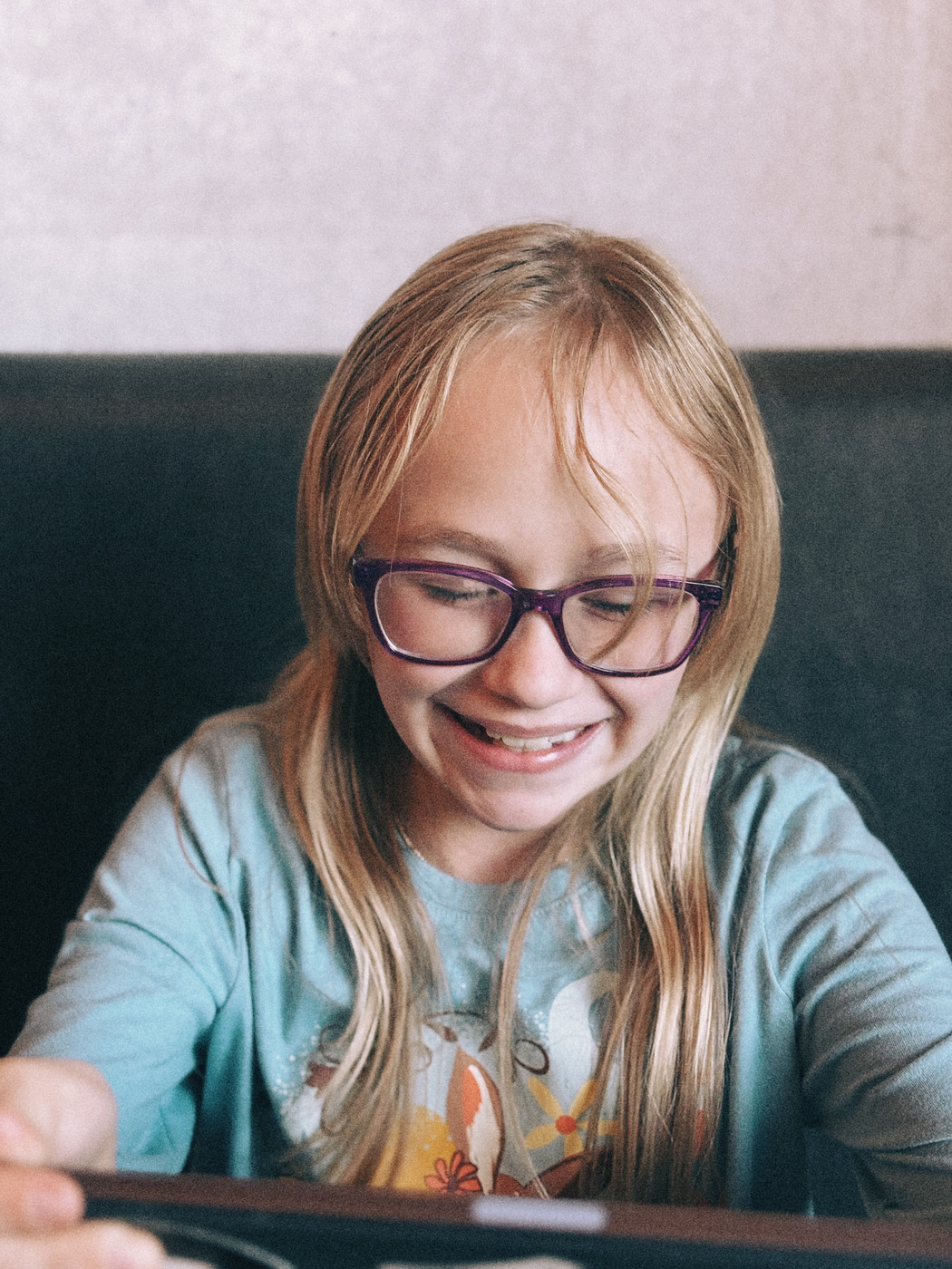 A young girl with long blond hair and purple glasses smiles brightly while looking down at a tablet. She’s wearing a light blue shirt with a colorful animal design, sitting against a dark seat and a softly lit wall in the background.