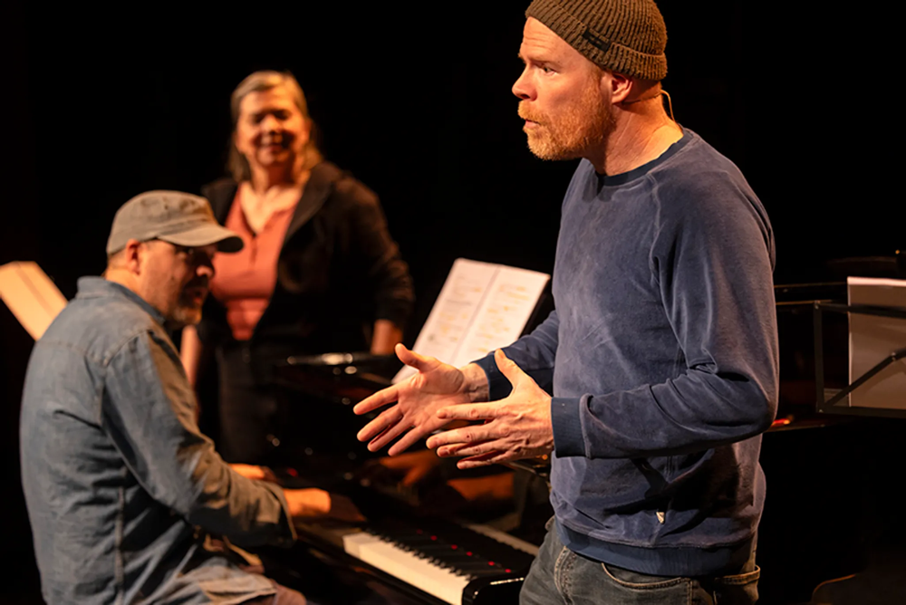 Three people on a dark stage: in the foreground a man wearing a brown beanie and a blue long-sleeve shirt gestures with his hands; to the left a pianist in a gray cap sits at a piano; in the background a smiling woman in a black jacket over an orange top stands near sheet music stands.