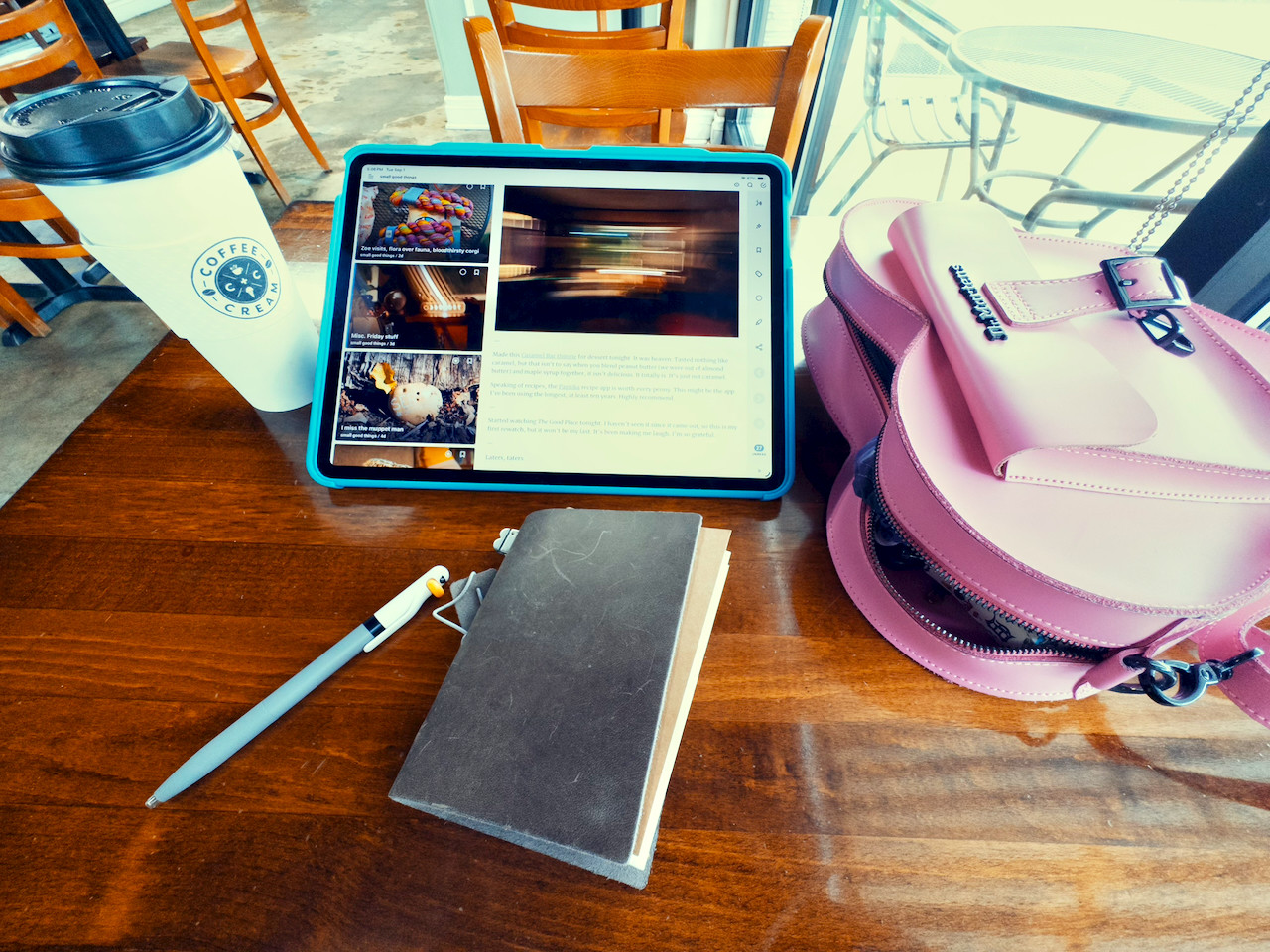 An over the top shot of a brown table, small leather notebook, iPad 13 inch, pink heart shaped handbag, and large coffee.