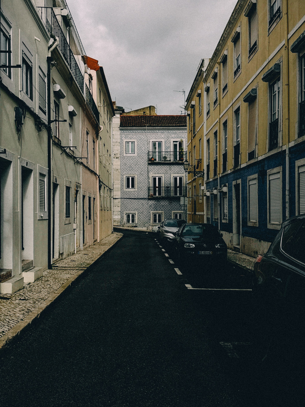 Narrow European street lined with parked cars and colorful old buildings, with a cloudy sky overhead.