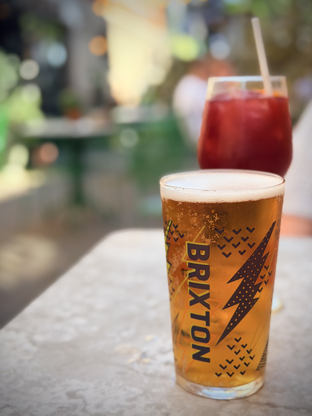 Pint of Brixton lager on a marble surface with a red iced cocktail with straw behind, in a blurred beer garden.