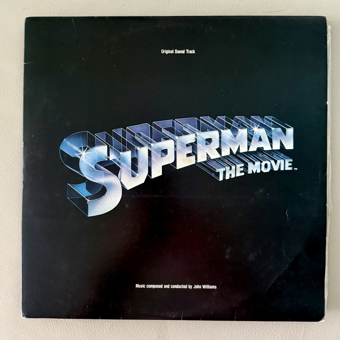 The front cover of the vinyl double album soundtrack to Superman The Movie (1978). The album is black with Superman The Movie curved across the front in large silvery blue all caps. At the top in smaller white lettering is “Orginal Sound Track” and across the bottom is “Music composed and conducted by John Williams”