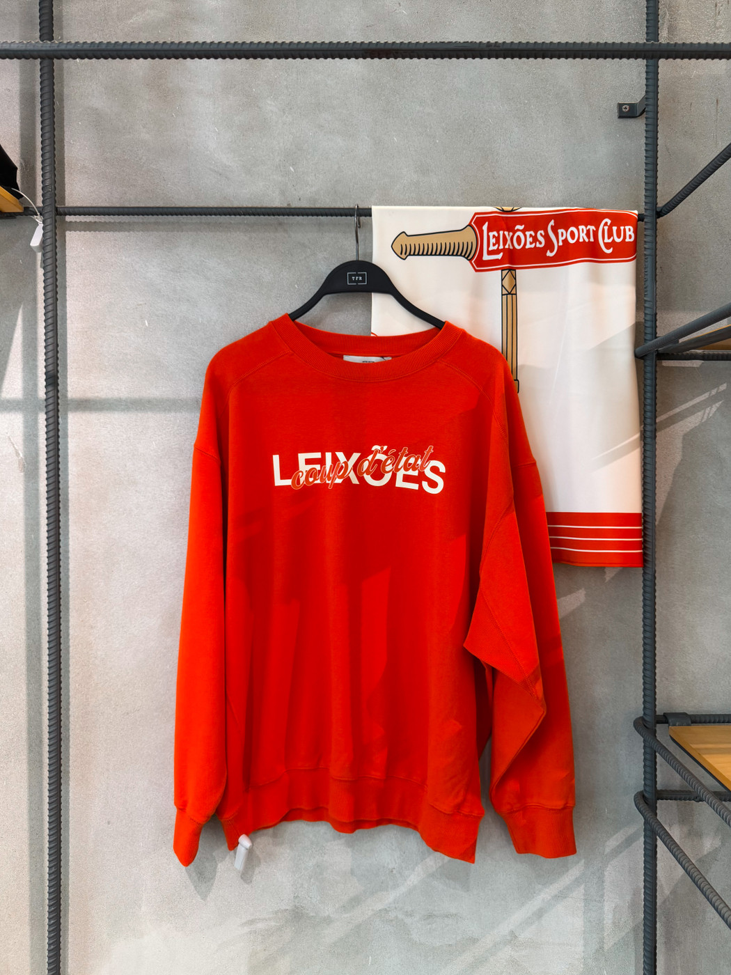 Red sweatshirt with the text “LEIXÕES coup d’état” hanging on a black hanger against a gray wall, with a Leixões Sport Club scarf partially visible in the background.