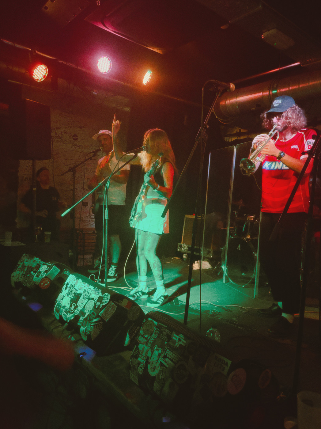 A small band performs on a dimly lit stage with red/orange lighting. On the right, a trumpet player with long hair, a blue cap, and red shirt; in the center, a blonde female guitarist/vocalist in a light dress sings into a microphone; to the left, a man wearing a hat stands near another mic. The front edge of the stage is lined with stickers and posters, and dark silhouettes of the audience are visible in the foreground.