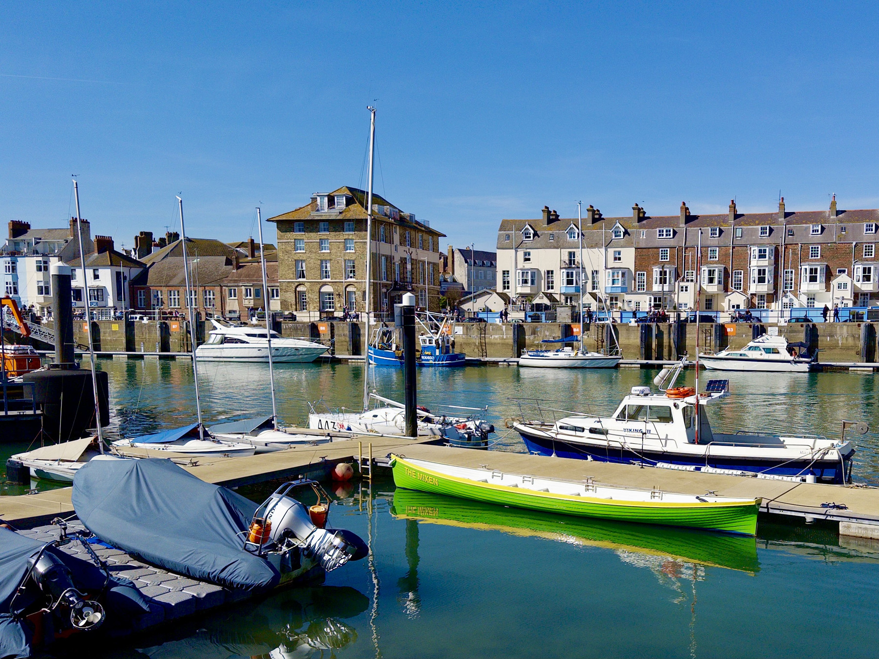 Small boats moored in a calm harbour with clear blue water, backed by a row of waterfront buildings under a bright sky.