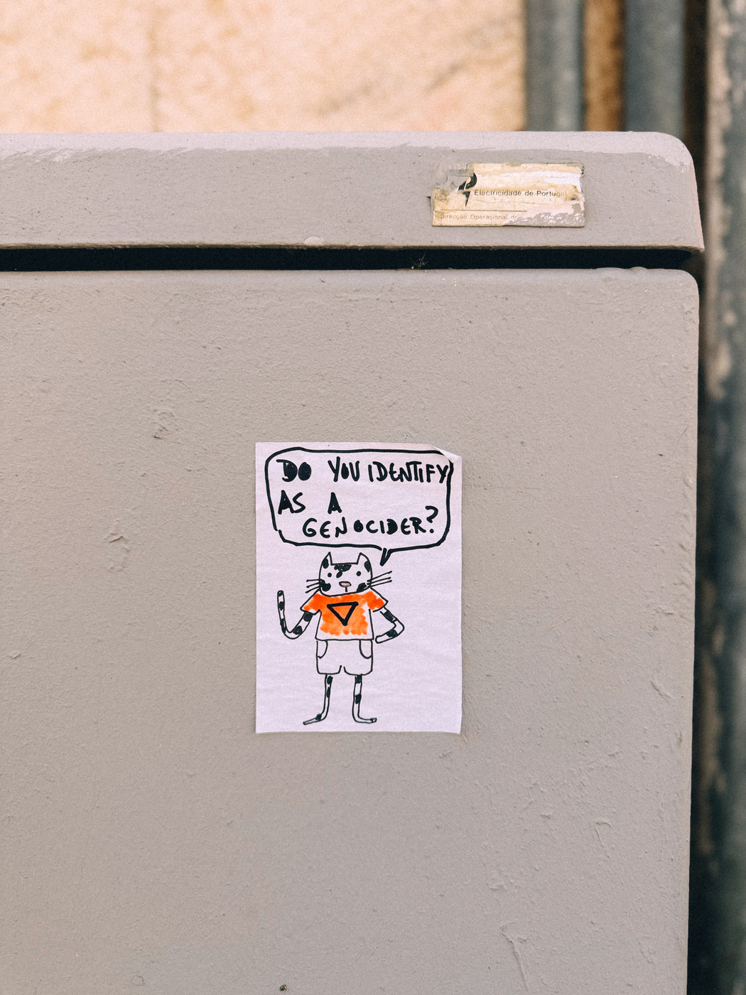 A hand-drawn poster of a cartoon cat wearing an orange shirt and shorts taped to a gray utility box, with a speech bubble saying, “Do you identify as a genocider?”