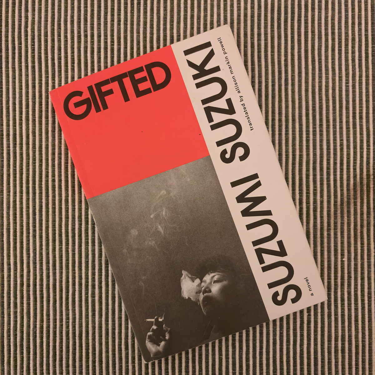 Paperback book titled Gifted by Suzumi Suzuki, shown at an angle on a striped fabric surface. The cover features bold black text on red and white blocks, with a black-and-white photograph of a woman smoking in the lower section.
