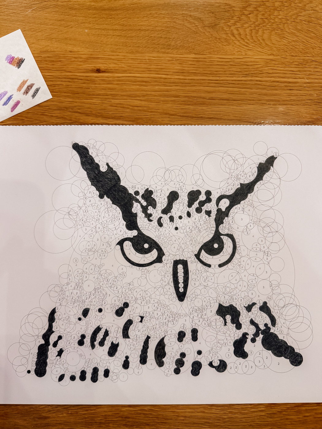 An intricate owl drawing is composed of numerous circles, partially colored in black, with a small paper showing color samples beside it.
