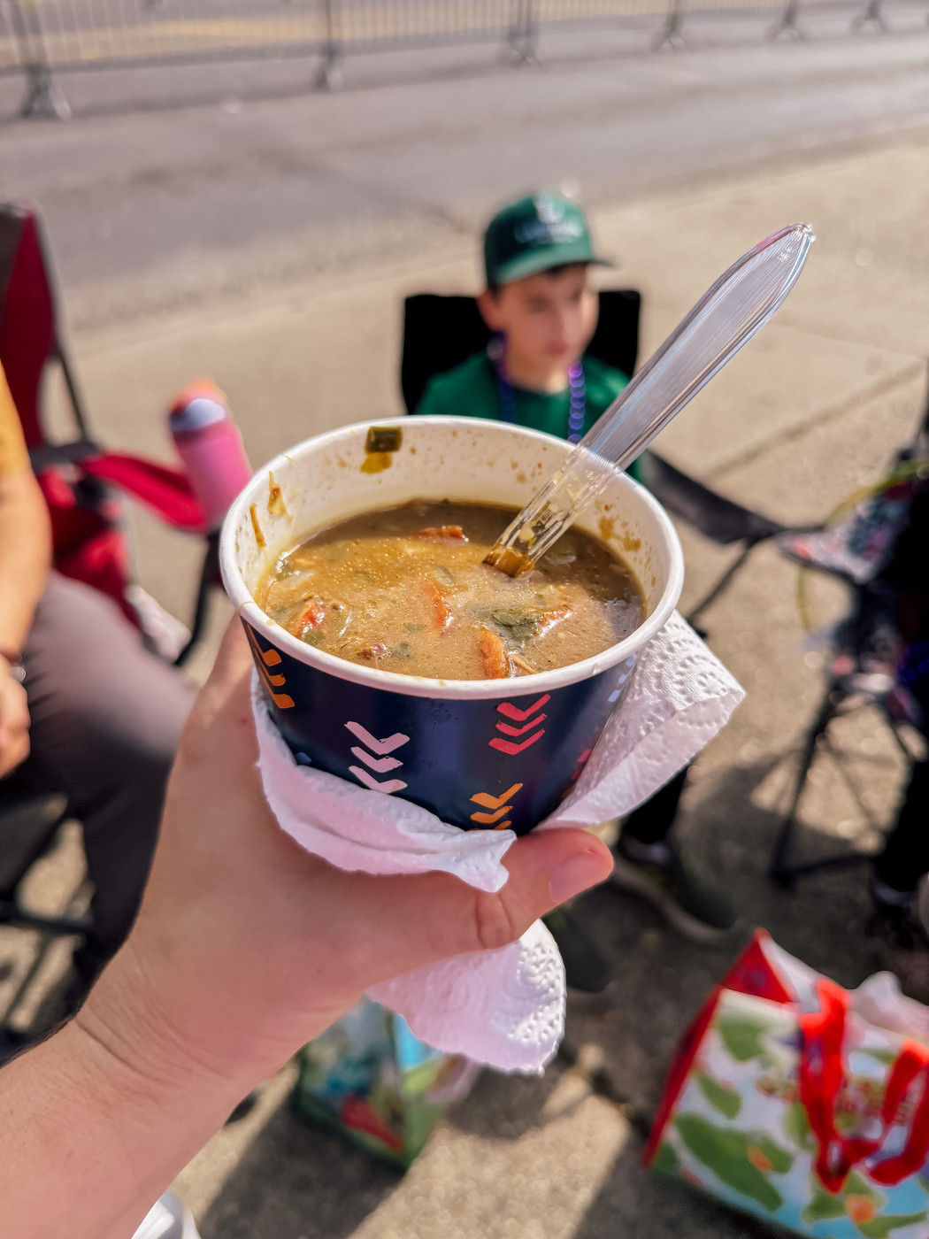A hand holding a cup of gumbo with a spoon in it, while a person sits blurred in the background.