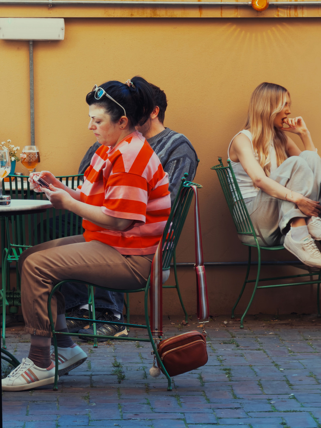 People seated at outdoor café tables against a yellow wall, with one person using a phone and another sitting turned away.