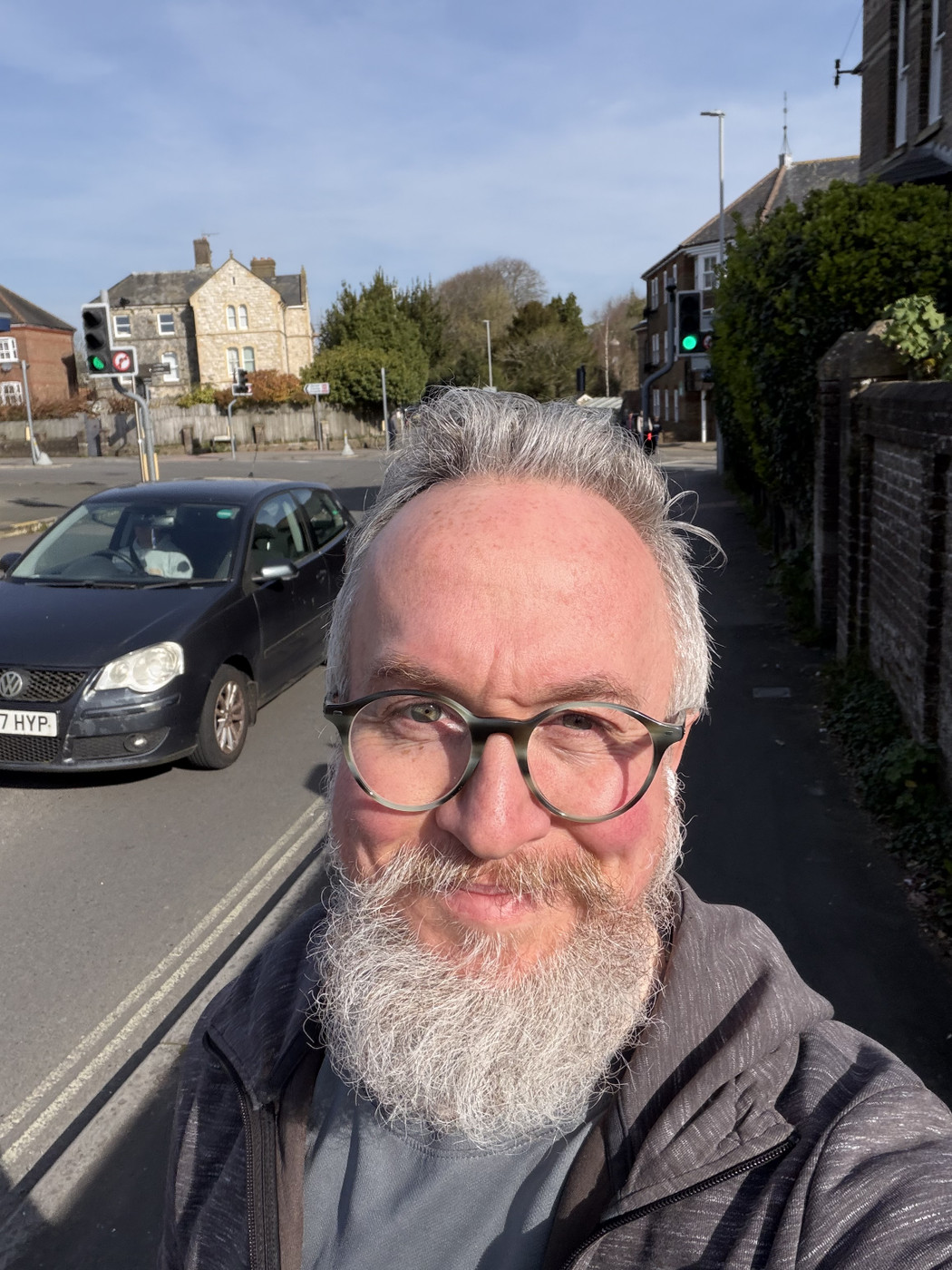 A smiling man with a white beard and glasses stands on a street corner. He is wearing a grey jacket and a blue shirt. Behind him is a black Volkswagen car and a row of houses. The sky is blue with some clouds. The mood of the image is cheerful and relaxed.