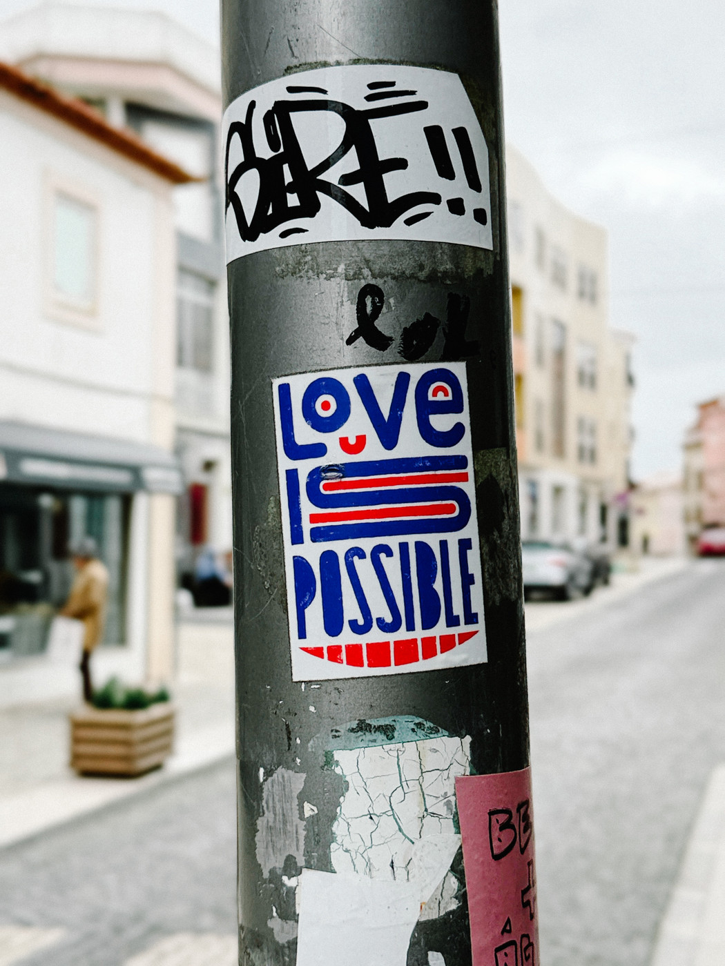A gray street pole covered with stickers, including a colorful one that says “LOVE IS POSSIBLE,” with a blurred urban street and parked cars in the background.