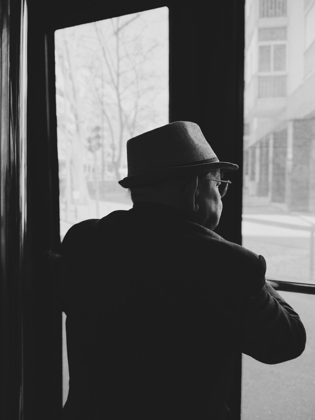 Black and white photo of an elderly man wearing a hat, looking out the window of a tram or bus toward a city street.