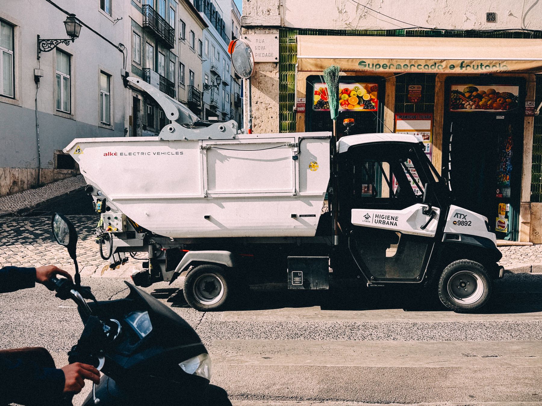 Small white electric urban hygiene truck parked on a street in front of a supermarket, with a person riding a scooter passing by.