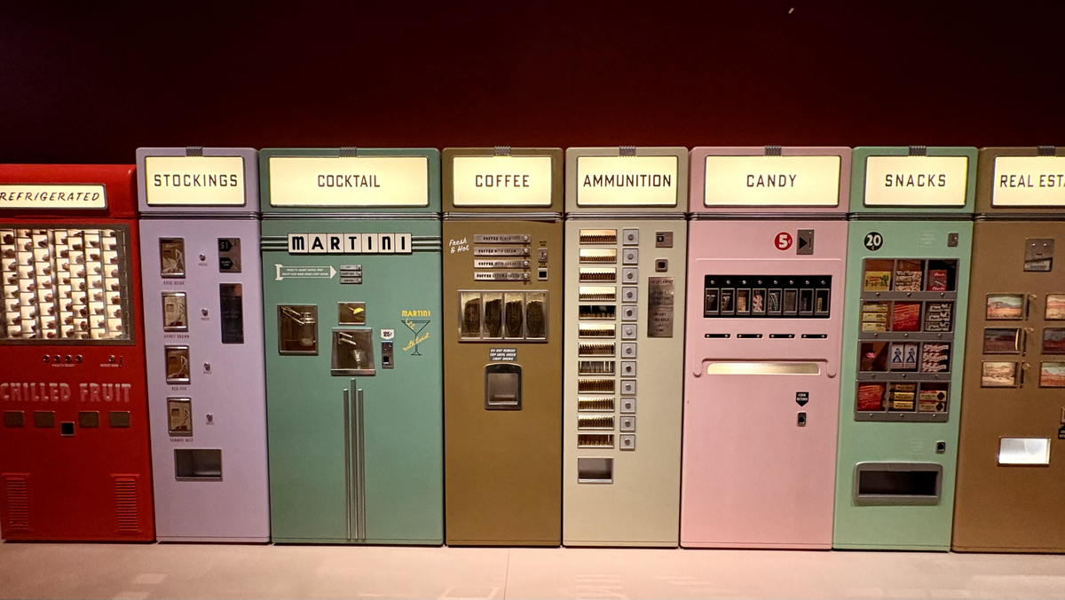 Row of brightly coloured vintage vending machines labelled with unusual categories such as ‘Stockings’, ‘Cocktail’, ‘Coffee’, ‘Ammunition’, ‘Candy’ and ‘Snacks’, displayed side by side against a dark red wall.