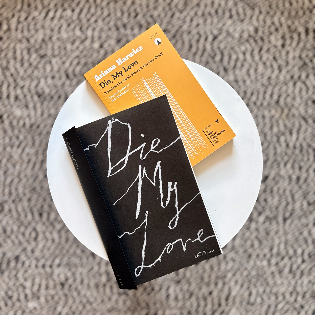 Two paperback books resting on a small round white table against a textured carpet, one with a bright orange cover titled Die, My Love by Ariana Harwicz and the other with a black cover reading Die My Love in large handwritten-style lettering.