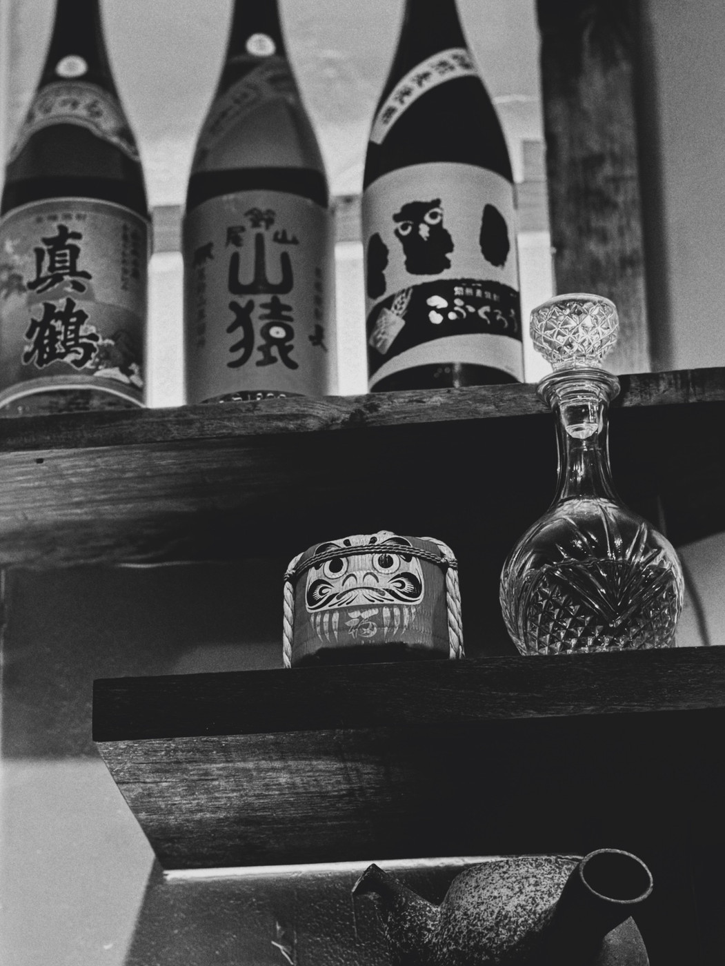 Black and white photo of a wooden shelf with Japanese sake bottles, a crystal decanter, a small Daruma doll, and a rustic teapot.