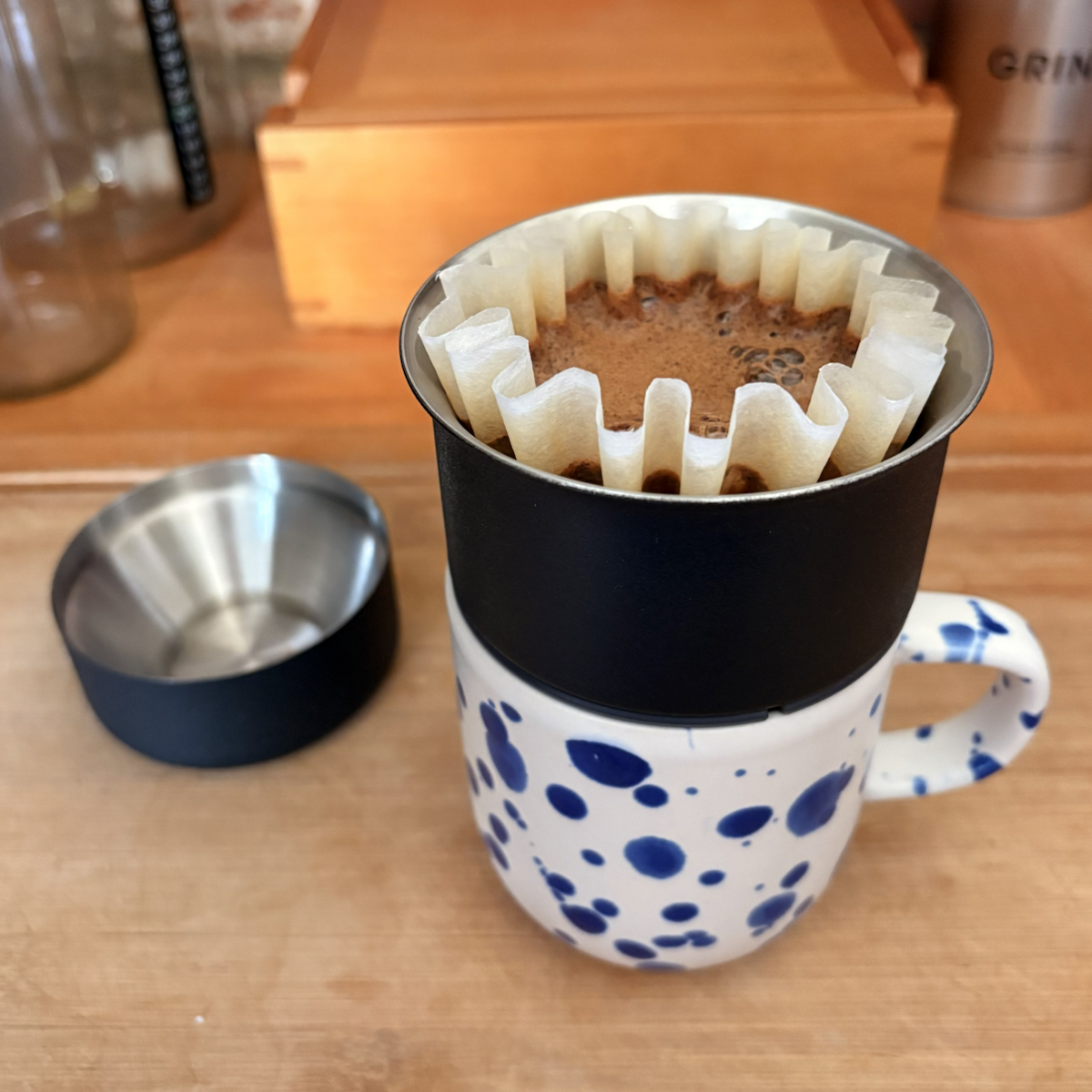Pour-over coffee brewing in a white ceramic mug with blue spots, using a paper filter set inside a black dripper. The mug sits on a wooden countertop beside a small metal cup, with coffee equipment blurred in the background.