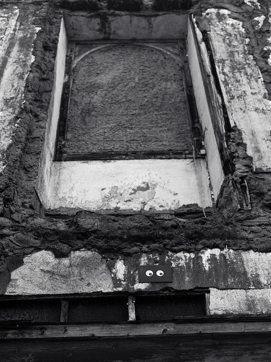 Black and white photo of a weathered, boarded-up window on a decaying building facade with a small black rectangle featuring two googly eyes attached below the window.