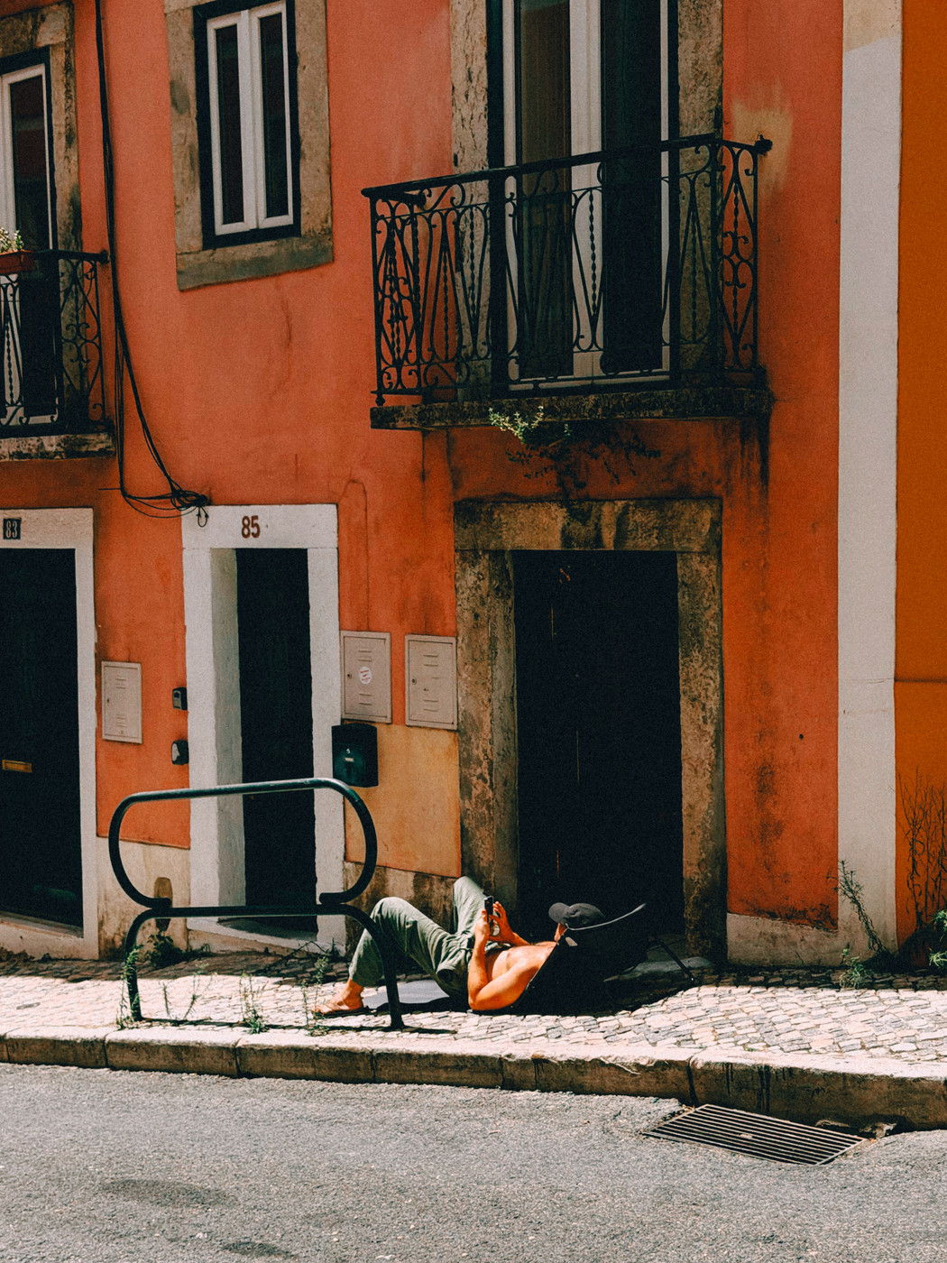 A person lies on a narrow cobblestone sidewalk in front of an orange building with balconies, sun-bathing , using a phone while partially shaded by a black cap.