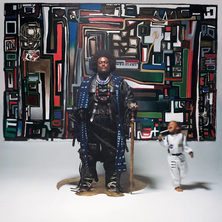 The album cover for Fearless Movement: A man stands centered in a striking portrait, wearing an elaborate indigo-dyed coat over layered leather garments adorned with colorful beaded necklaces and ornamental accessories. He has a large natural afro and holds a tall staff, standing stoically on a circular platform. Behind him hangs a large abstract painting filled with geometric shapes in red, blue, white, black, and earth tones, evoking a modernist or Afrofuturist aesthetic. To his right, a young child in a light gray outfit is captured mid-motion, slightly blurred, appearing to run or dance joyfully. The setting is a clean studio space with a neutral floor, creating a contrast between the richly detailed central figure and the dynamic energy of the child.