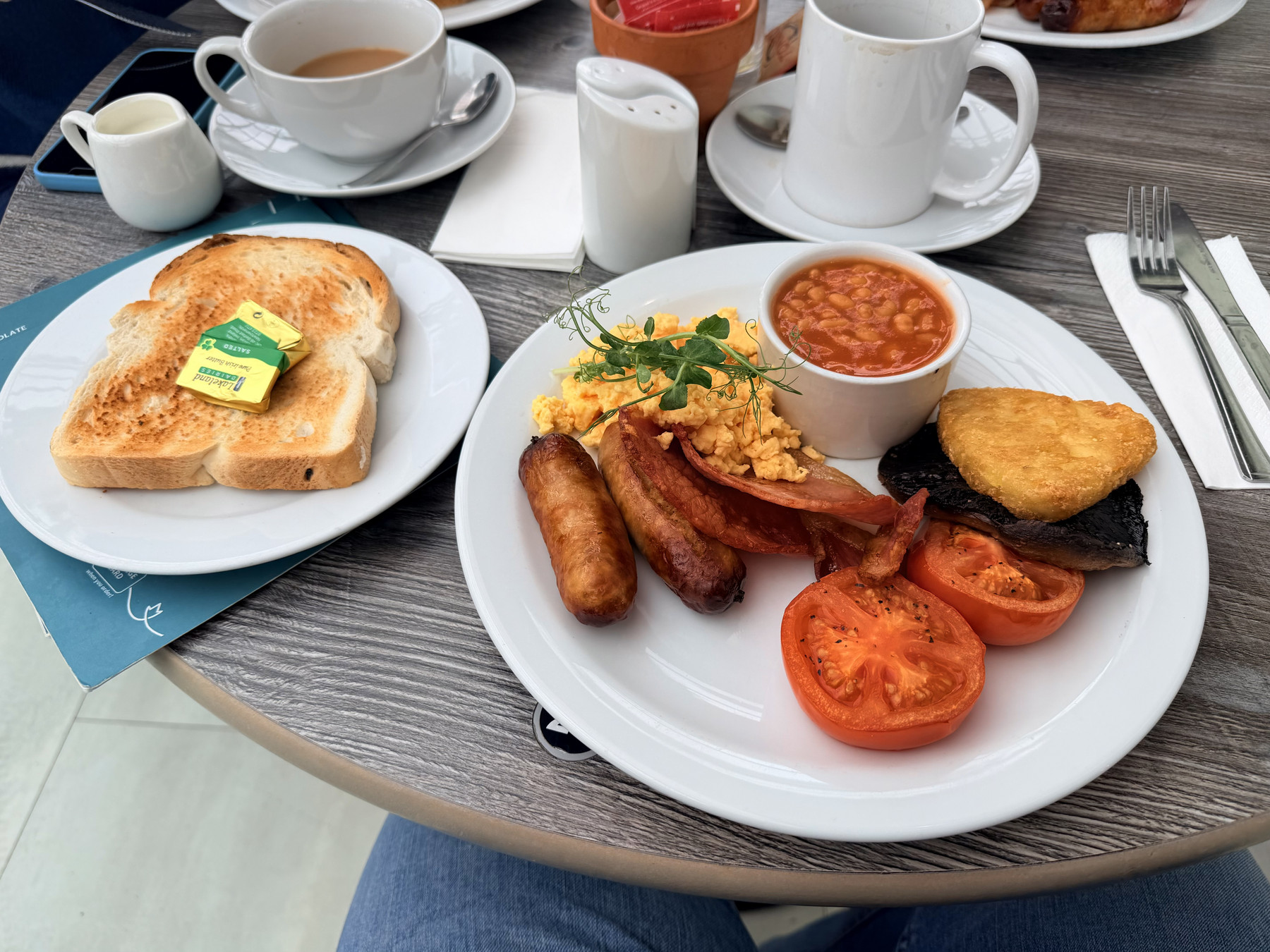 Two plates of breakfast food on a wooden table. One plate has toast with butter and a cup of coffee. The other plate has scrambled eggs, sausages, grilled tomatoes, mushroom, baked beans, and hash brown. The mood is warm and inviting, suggesting a hearty morning meal.