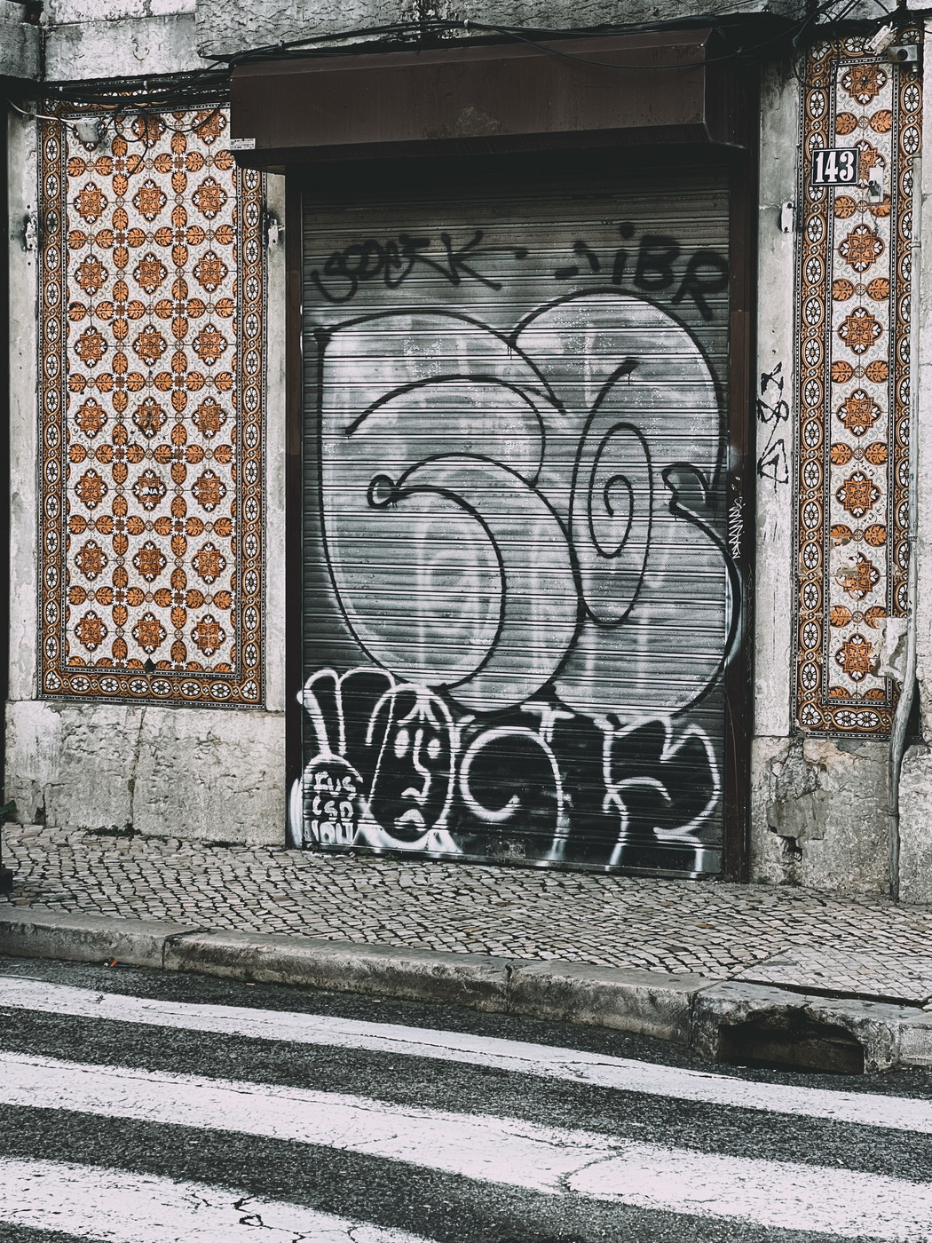 A street scene featuring a closed metal shutter door with graffiti, framed by traditional patterned tiles. The ground is cobblestone, and there’s a pedestrian crossing in the foreground. The building number “143” is visible.