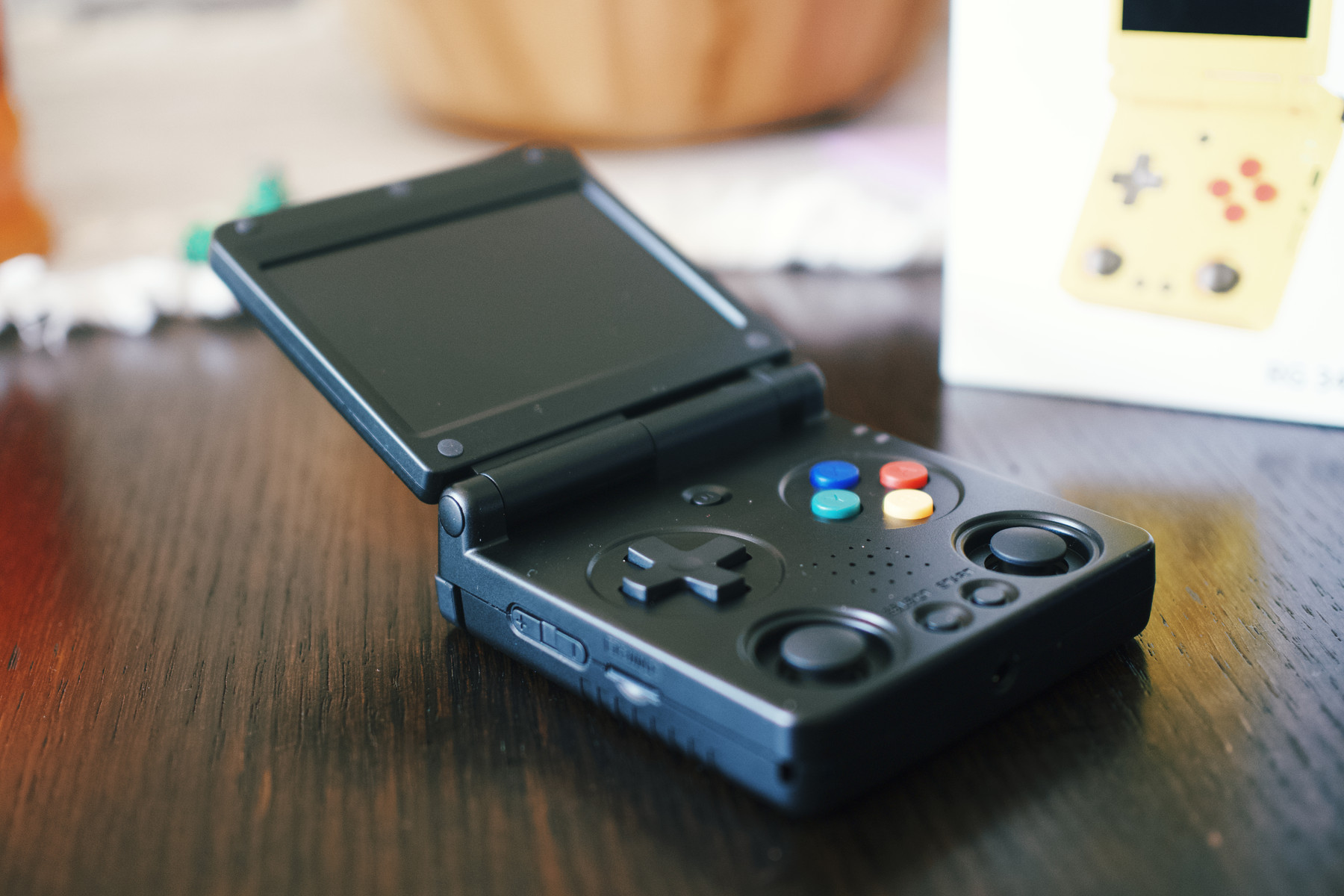 A black handheld retro-style gaming console with a clamshell design, open to reveal its screen and colorful control buttons. The device sits on a wooden table, with a blurred yellow version of the same console visible in the background.